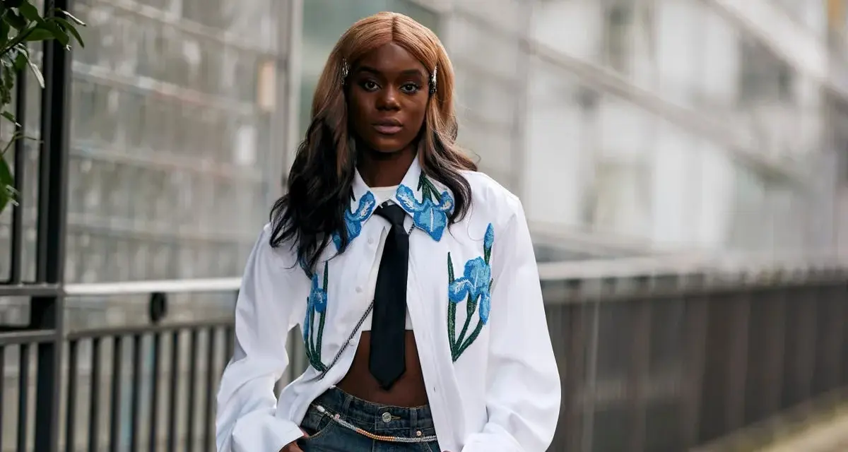 Modella con jeans baggy, un capo che simboleggia libertà e stile informale, indossa una camicia bianca con ricami floreali e cravatta.