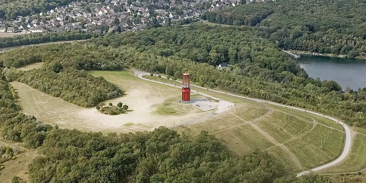 Luftaufnahme der Halde Rheinpreußen mit rotem Geleucht, umgeben von Wald und einem See.