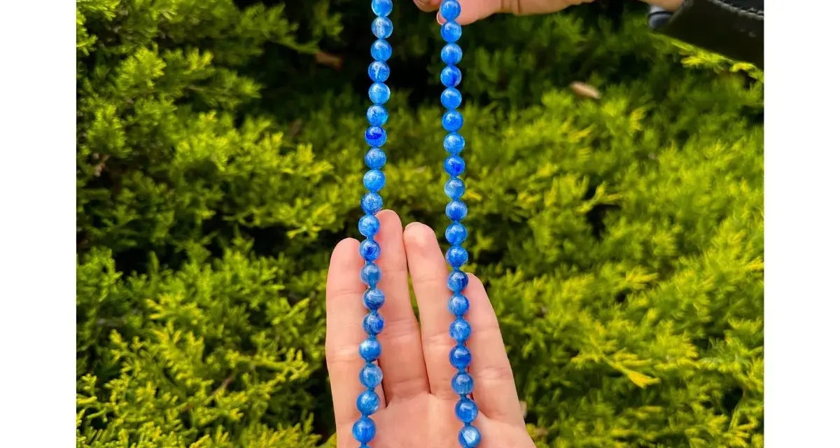 Collana di cianite blu, con perle luminose che catturano la luce, tenuta tra le mani su uno sfondo verde.