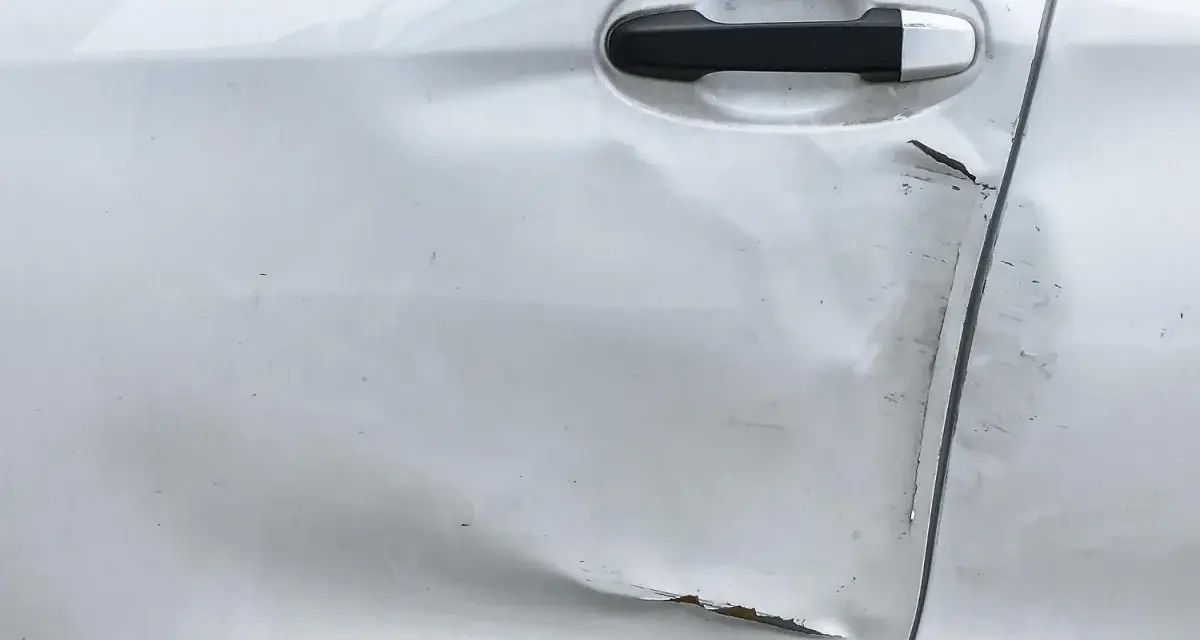 Widoczne wgniecenie i zarysowania na srebrnych drzwiach samochodu, świadczące o uderzeniu drzwiami na parkingu.