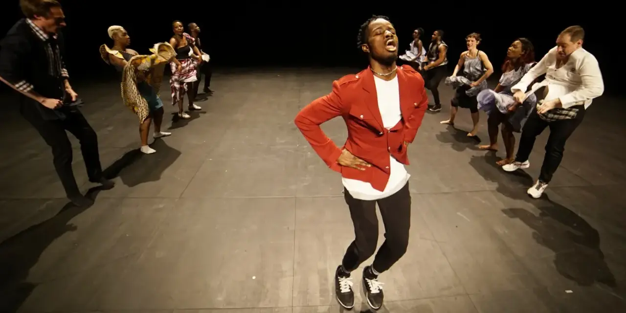 Un danseur afro 93, en veste rouge, mène une troupe sur scène. Les danseurs, aux tenues variées, exécutent des mouvements synchronisés.