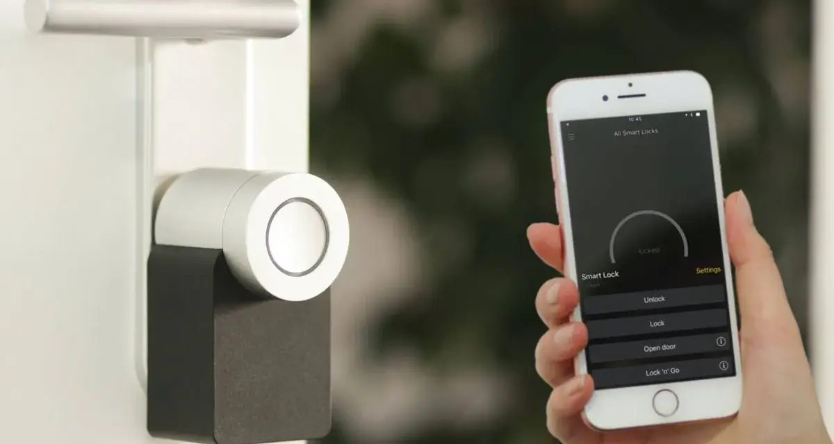 Mano sostiene un iPhone que muestra una app para controlar un cerrojo inteligente. La puerta tiene un diseño moderno.