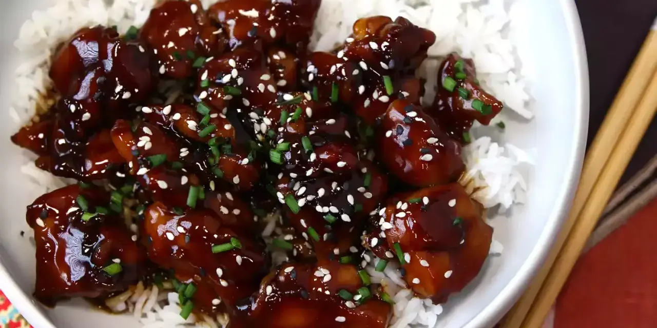 Trozos de pollo teriyaki glaseados y brillantes, cubiertos con semillas de sésamo y cebollino, servidos sobre una cama de arroz blanco.