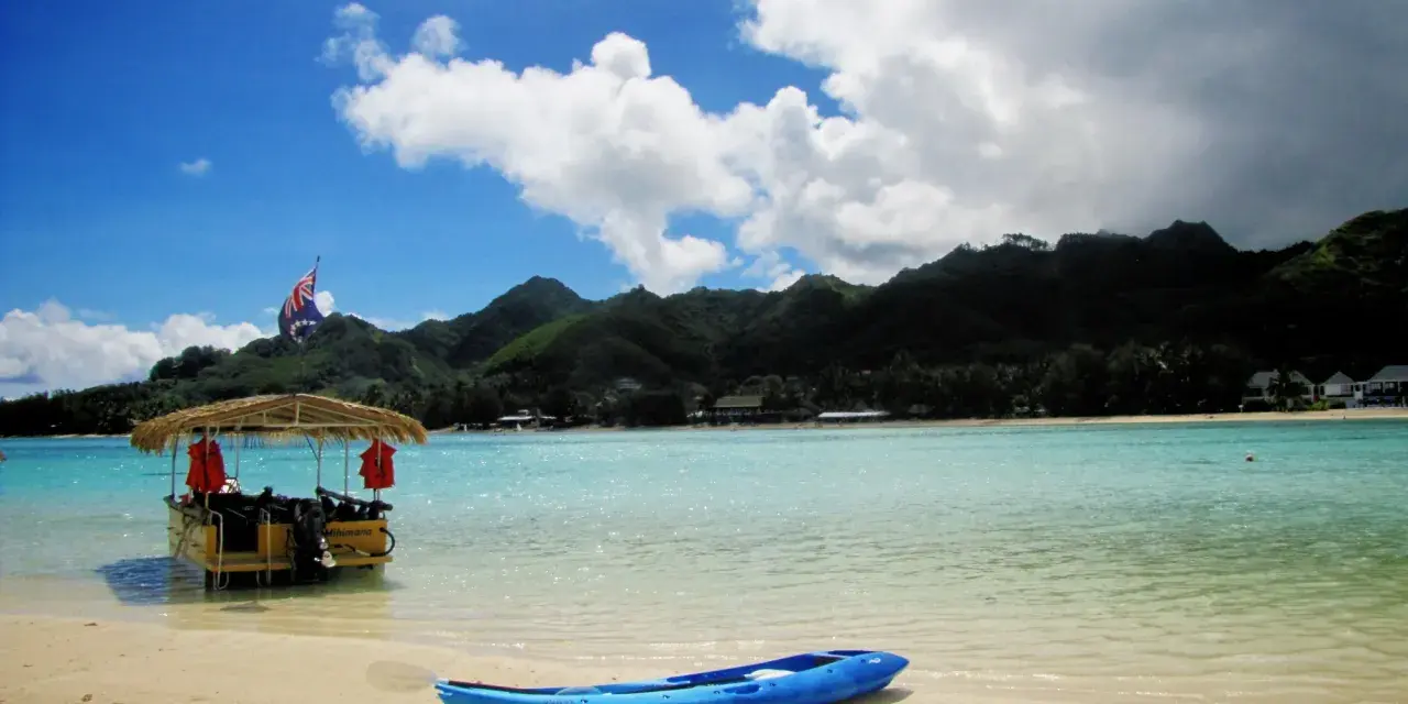 A blue kayak rests on a sandy beach in the Cook Islands, with a thatched-roof boat and lush green mountains in the background.