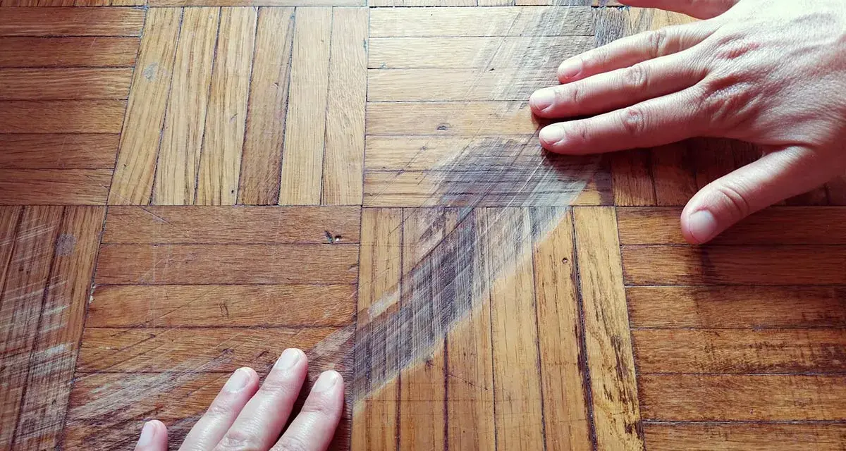 Mani sul parquet rovinato, mostrando i graffi e le macchie. Si valuta come pulire il parquet rovinato per riportarlo al suo antico splendore.