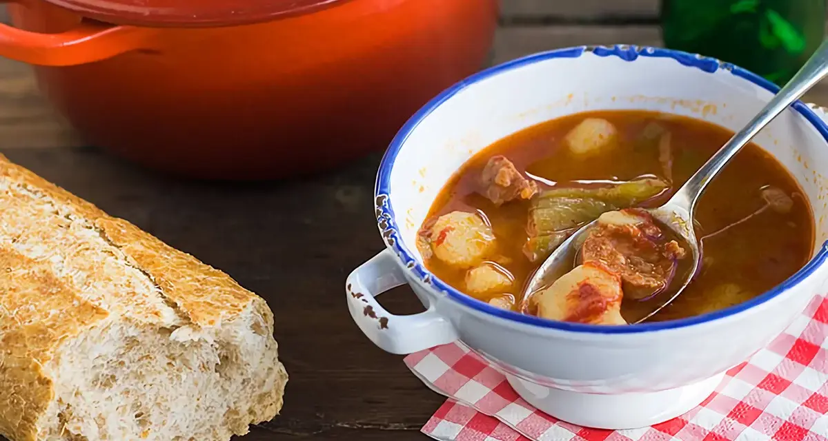Un reconfortante plato de patatas a la riojana, con chorizo y pimientos, servido en un cuenco blanco con borde azul, junto a una hogaza de pan rústico.