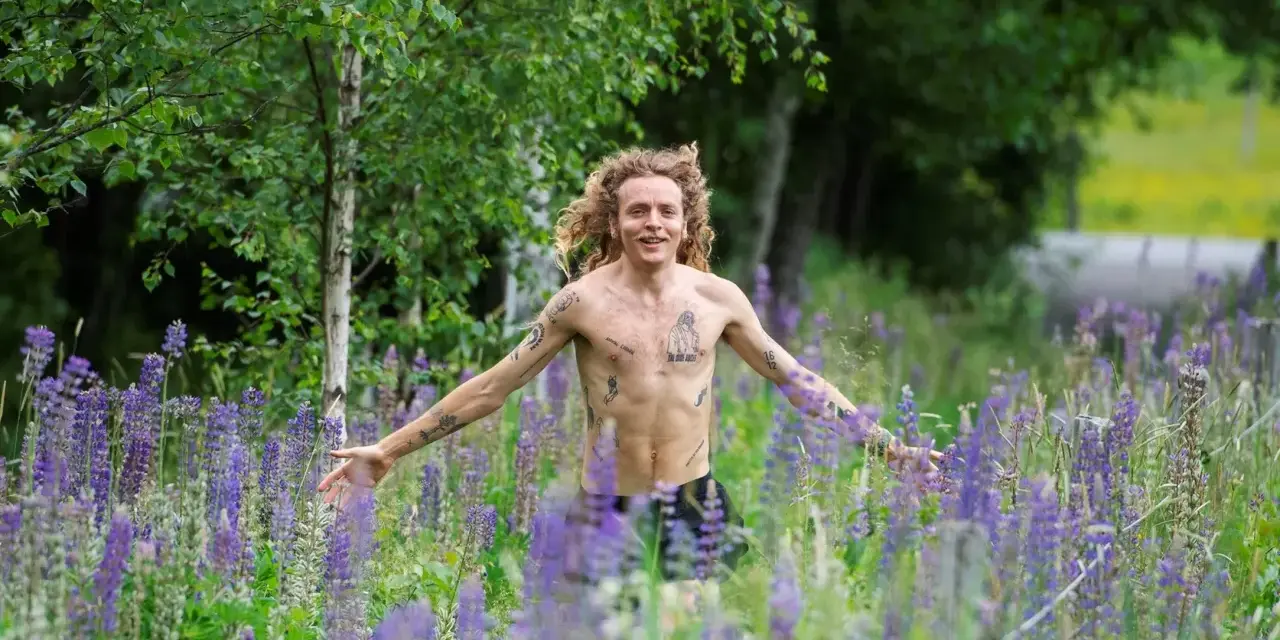 En man med lockigt hår och tatueringar står i en äng full av lila lupiner. Han ser ut att njuta av naturen, kanske som en del av ett avsnitt av "spring snyggt podden".