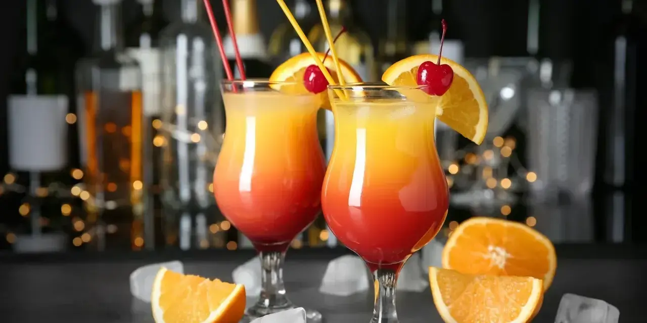 Dwa drinki Malibu z sokiem pomarańczowym, ozdobione pomarańczą i wiśnią, na tle baru.
