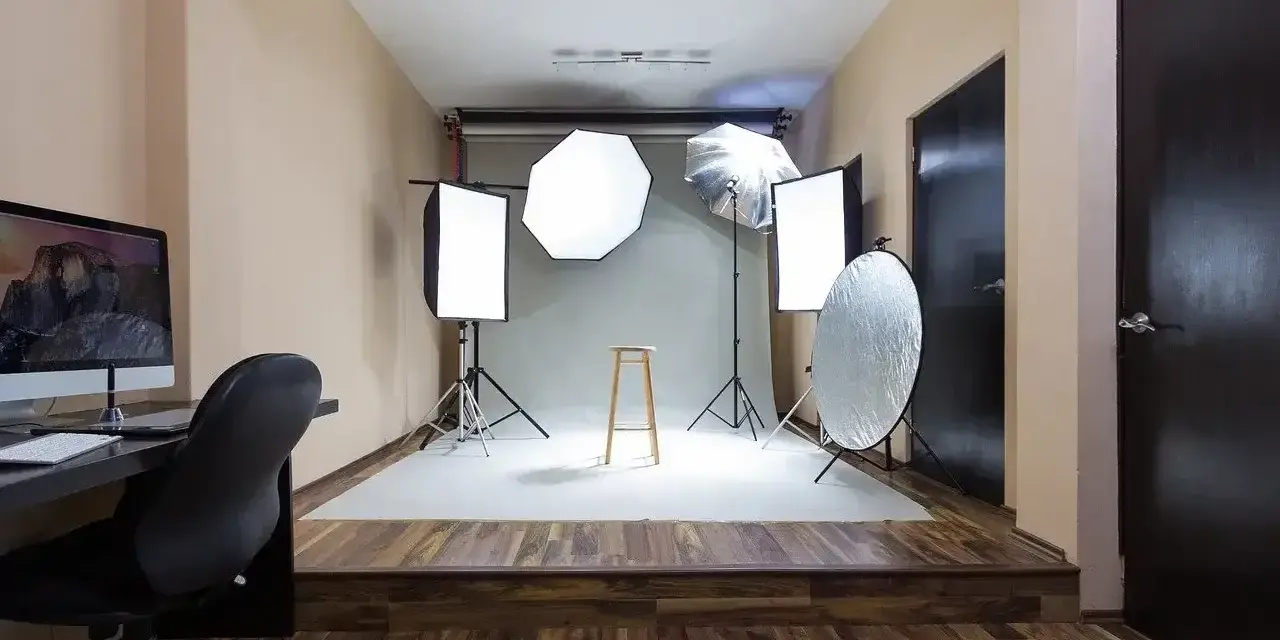 Domowe studio fotograficzne z oświetleniem, statywami, blendą i krzesłem. Po lewej komputer z monitorem.