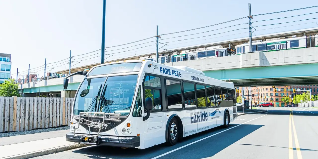 A white EZRide bus, offering fare-free transit from Boston to Cambridge, MA, drives past an elevated train station.