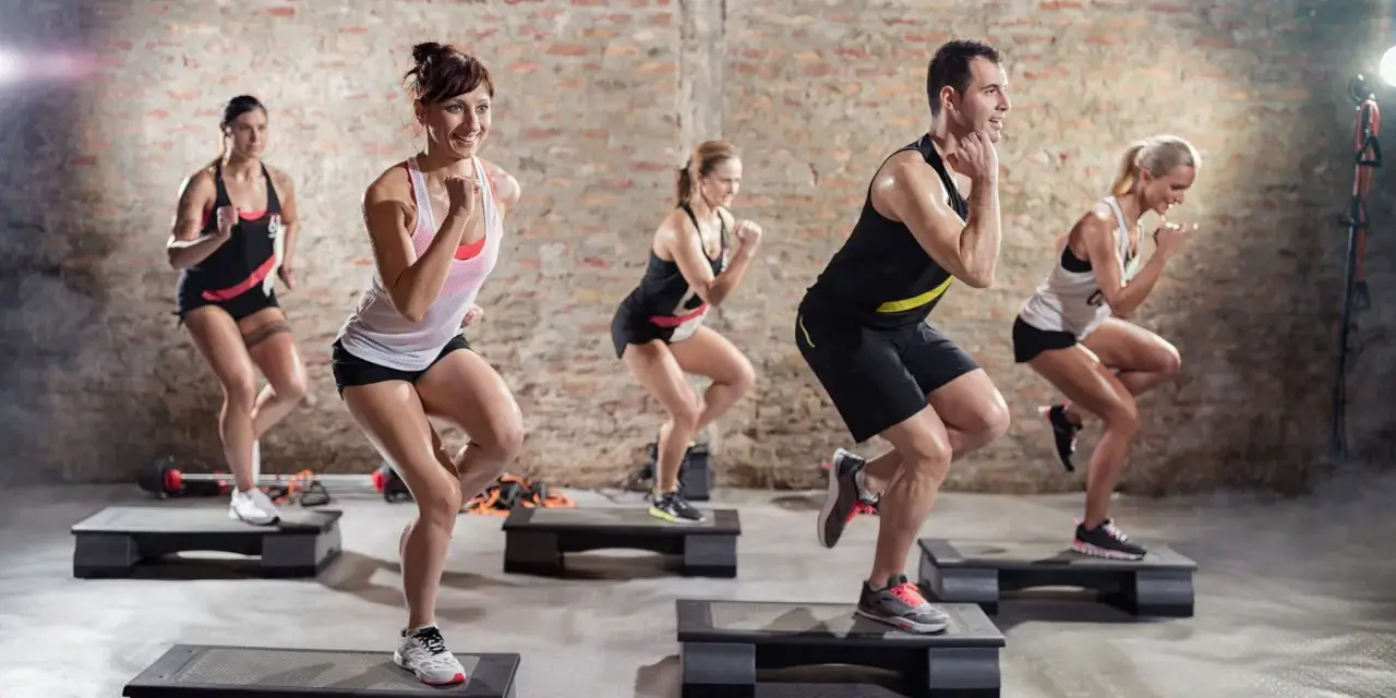 Un groupe de personnes participe à un cours step, effectuant des mouvements dynamiques sur des plateformes d'exercice dans une salle de sport.