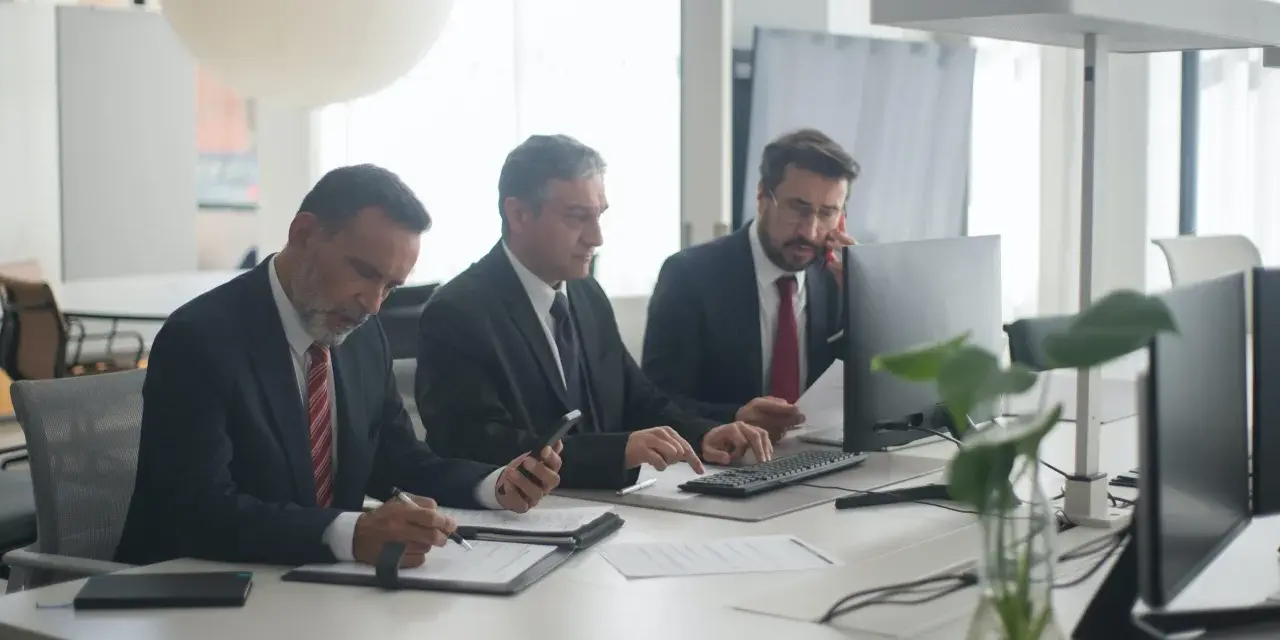 Trzech mężczyzn w garniturach przy biurku. Jeden pisze, drugi patrzy na telefon, trzeci rozmawia przez telefon. Czyżby to byli best s.a. oszuści?