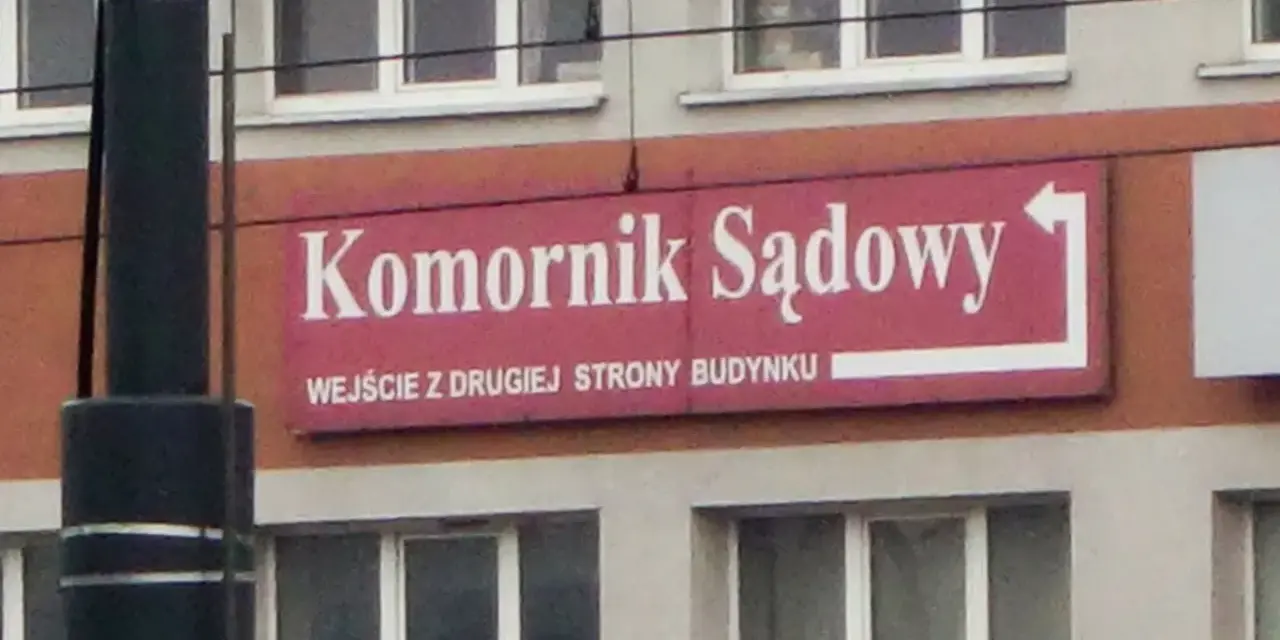 Tablica "Komornik Sądowy" na budynku. Wskazuje wejście z drugiej strony.