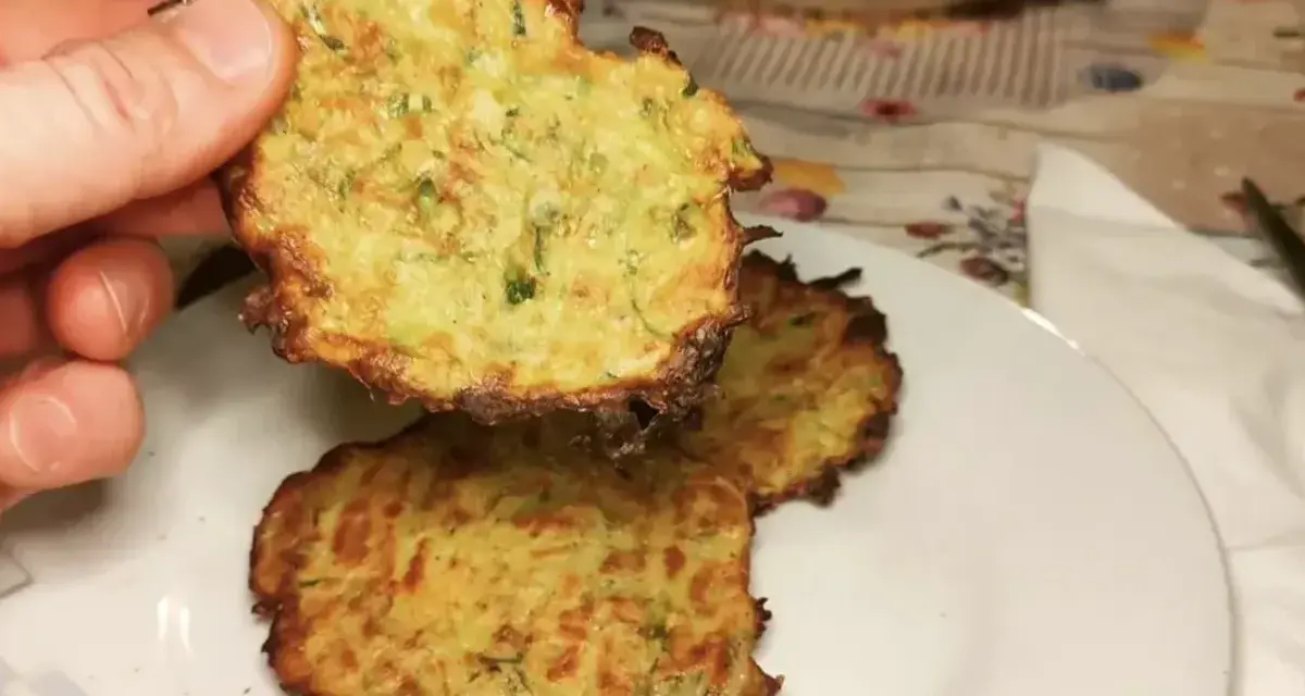 Una mano tiene una frittella di zucchine dorata e croccante. Sul piatto, altre frittelle di zucchine invitano ad essere gustate.