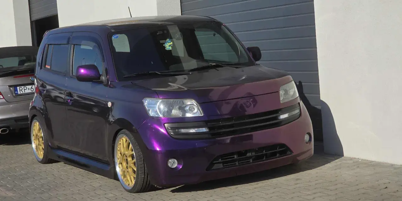 Fioletowy Daihatsu Materia z błyszczącymi złotymi felgami, zaparkowany przed garażem.