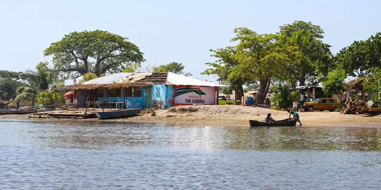 Nad rzeką w Gambii, przy brzegu z łodziami i budynkami, widać tablicę "Welcome to THE GAMBIA".