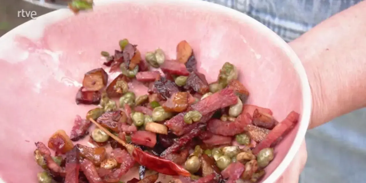 Un plato rosa lleno de habas con jamón, guisantes y un chile rojo seco.