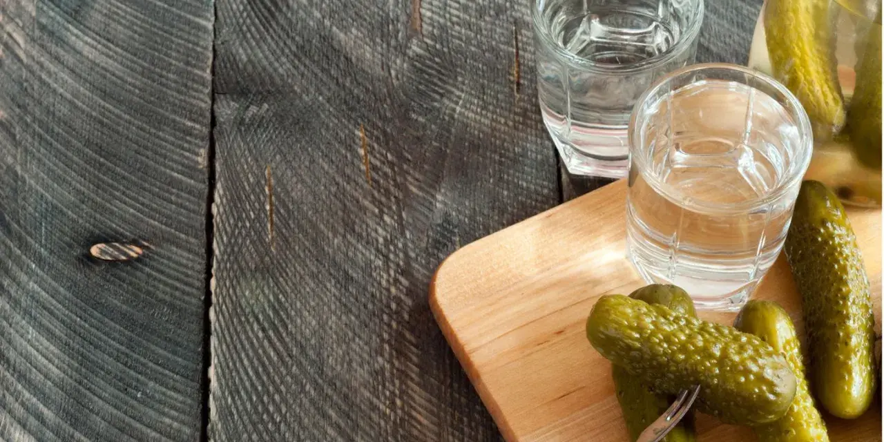 Zwei Gläser Wodka und eingelegte Gurken auf einem Holzbrett. Perfekt für alle, die Wodka selber machen wollen.