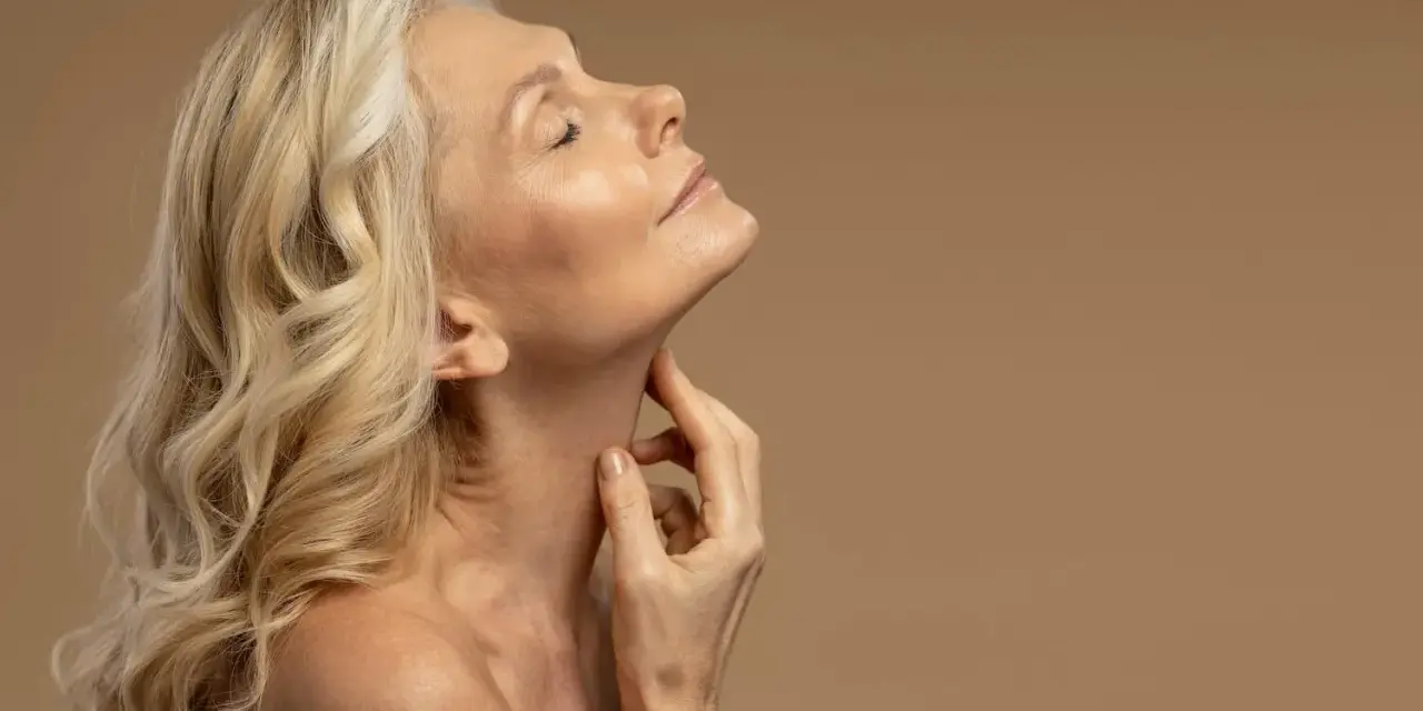 Kobieta z blond włosami i zamkniętymi oczami delikatnie dotyka szyi. Jej skóra w okresie menopauzy jest widoczna, podkreślając naturalne piękno.