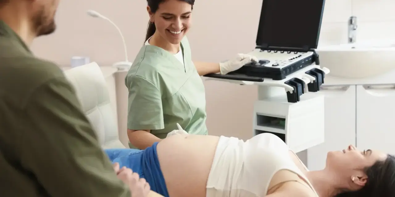 Uśmiechnięta lekarka wykonuje badanie wolnego płodowego dna u ciężarnej pacjentki, podczas gdy partner wspiera ją, trzymając za rękę.