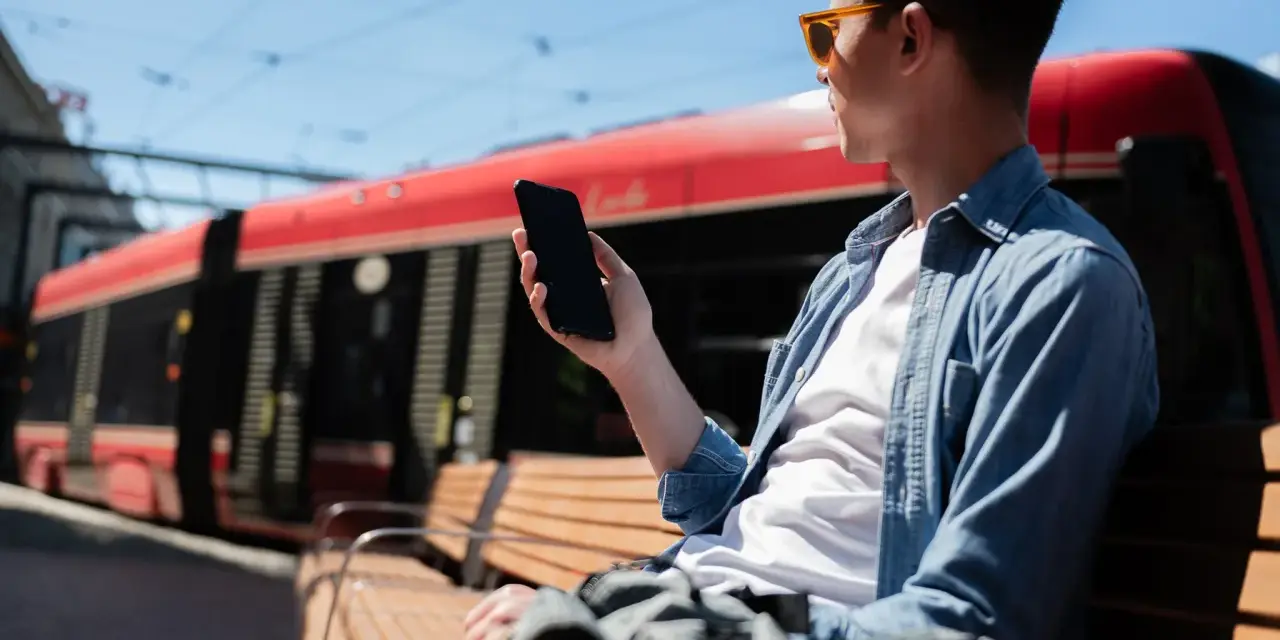 Mężczyzna w okularach siedzi na ławce i trzyma telefon. W tle czerwony tramwaj.