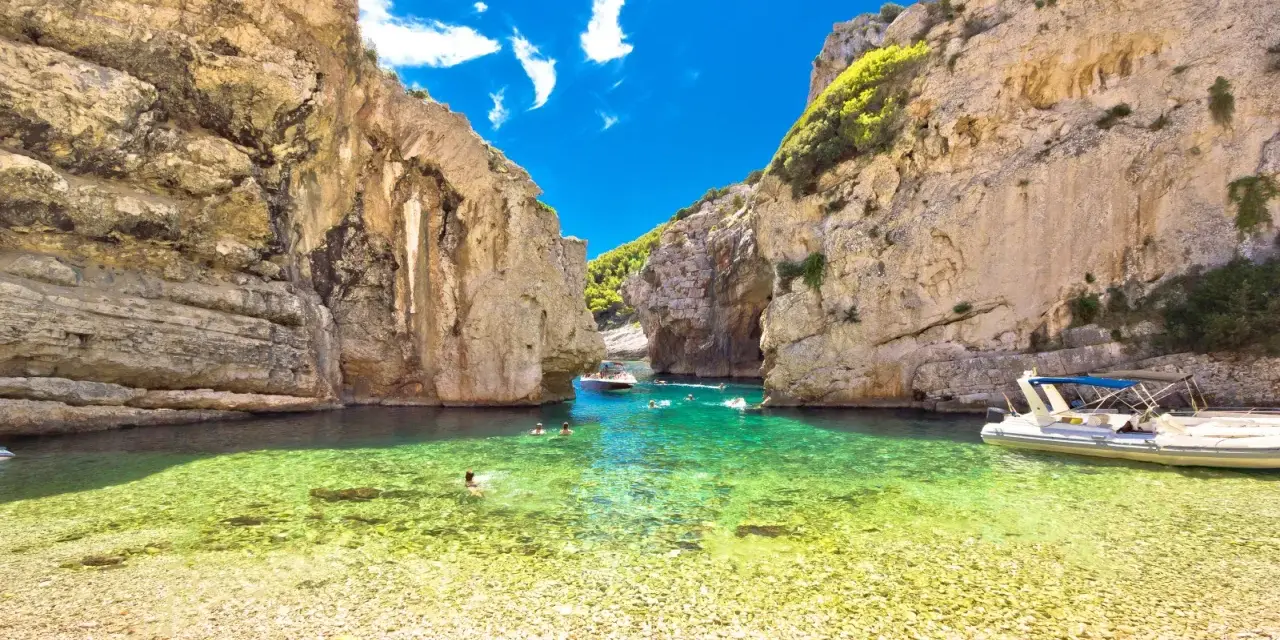 Spiaggia di ciottoli e acque cristalline tra le rocce dell'isola di Lissa, con persone che nuotano e barche.
