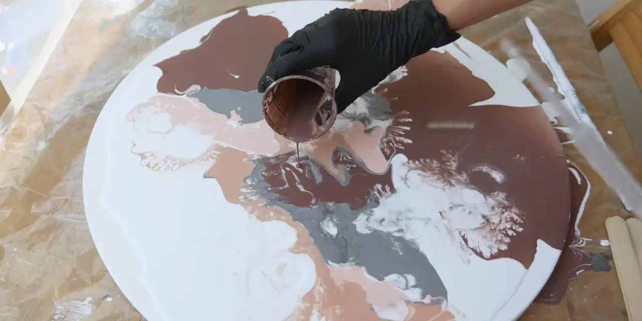 Main brune versée sur un tableau pouring acrylique blanc, créant des motifs abstraits.