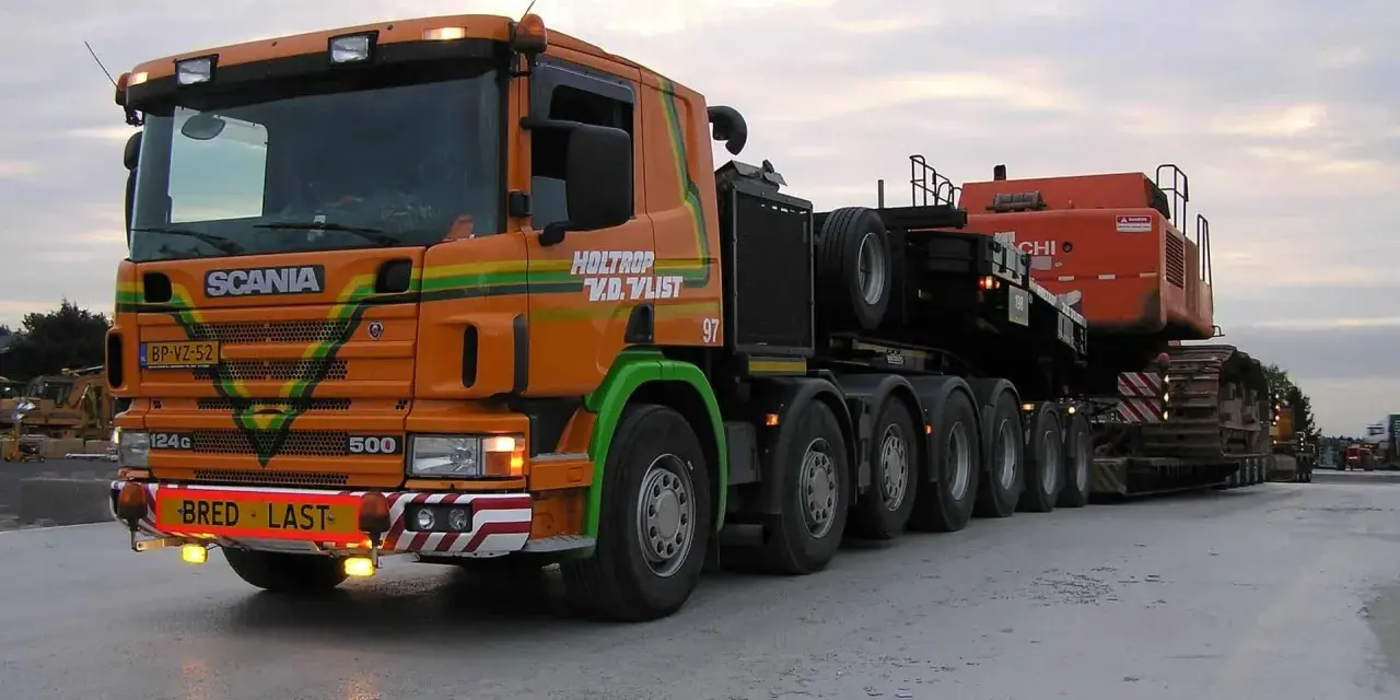 Pomarańczowy Scania z naczepą przewozi koparkę. Transport ponadgabarytowy wymaga specjalnych godzin jazdy.