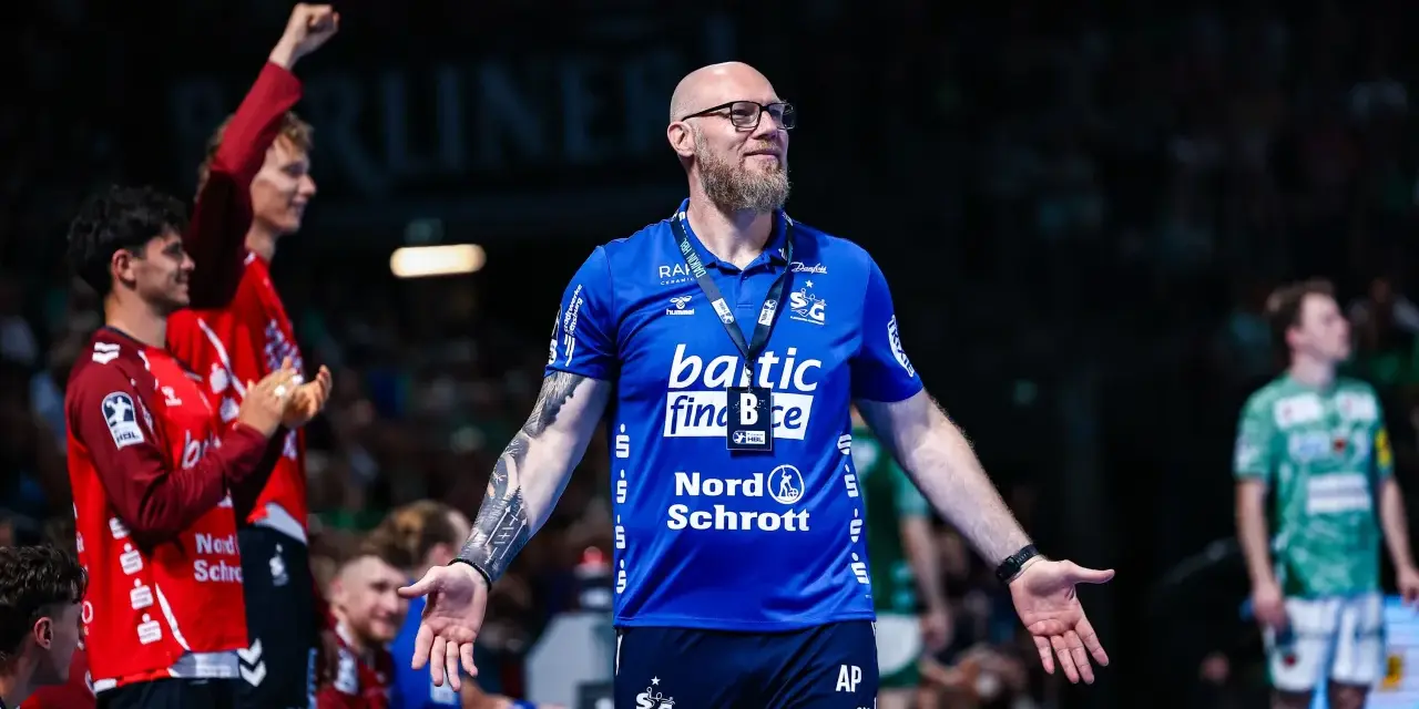 Trener SG Flensburg-Handewitt w niebieskiej koszulce z tatuażem na ramieniu, z otwartymi dłońmi, podczas meczu.