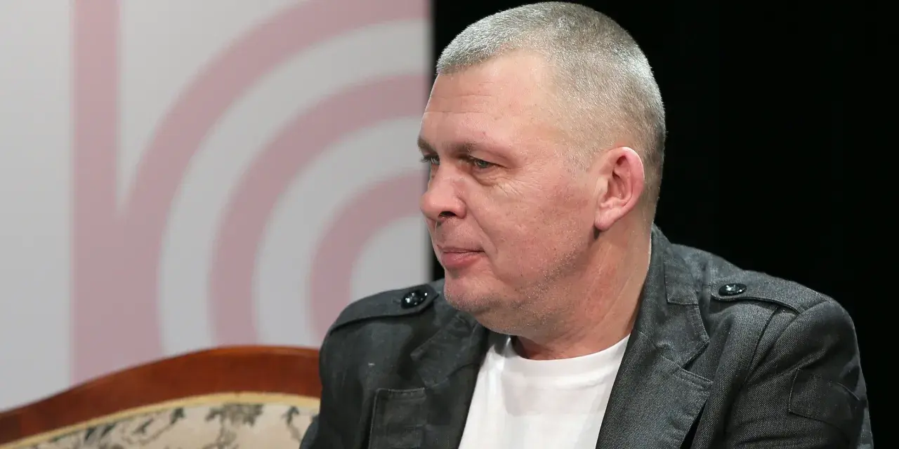 Adam z zespołu Raz Dwa Trzy w szarej marynarce, siedzi na kanapie.