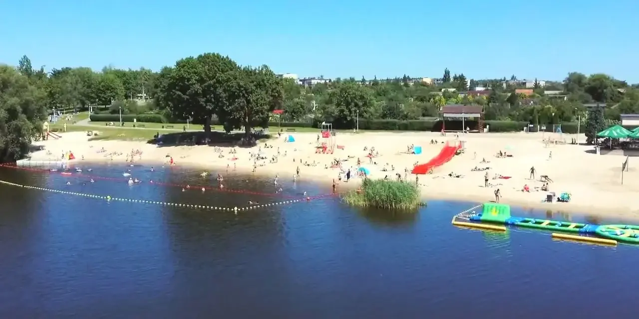 Słoneczny dzień nad jeziorem Grzymisławskim. Ludzie odpoczywają na plaży, kąpią się i bawią na dmuchanych atrakcjach.