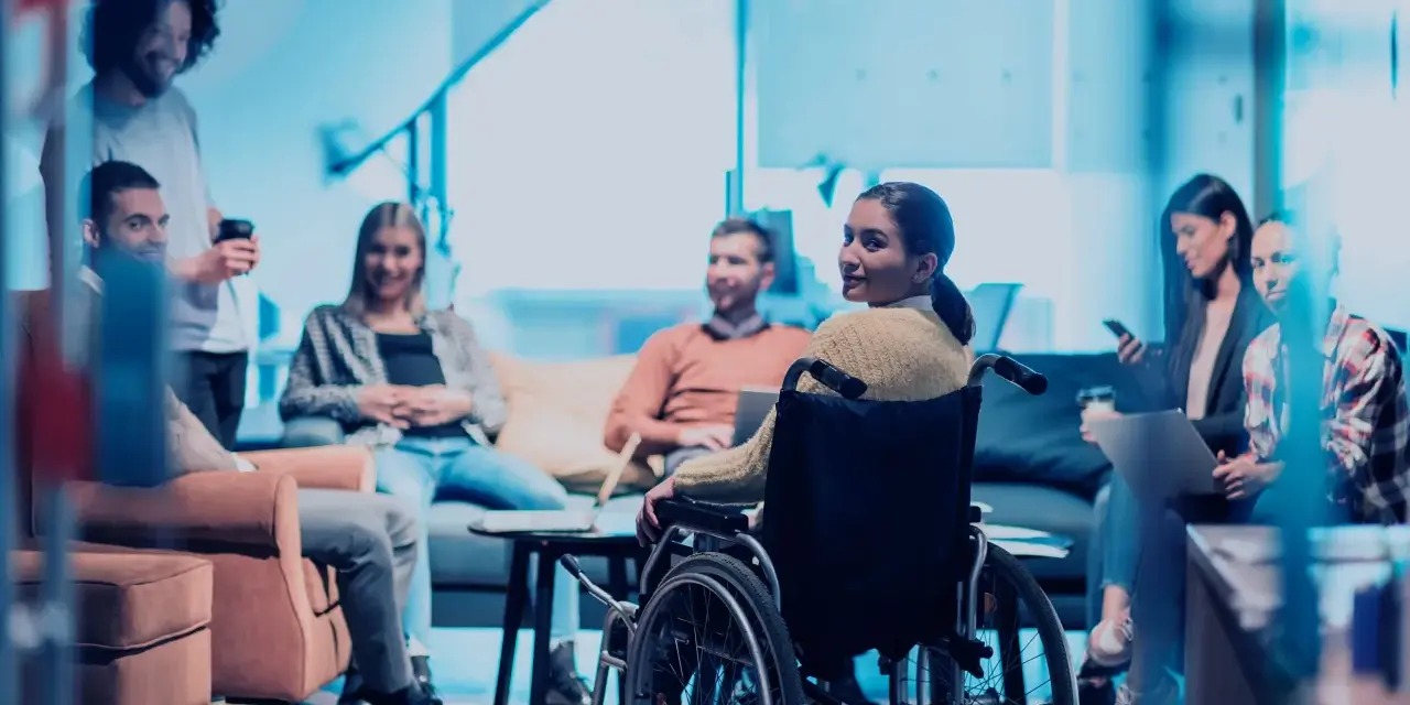 Ein Team bespricht sich, eine Frau im Rollstuhl ist integriert. Ein leidensgerechter Arbeitsplatz, an dem alle willkommen sind.