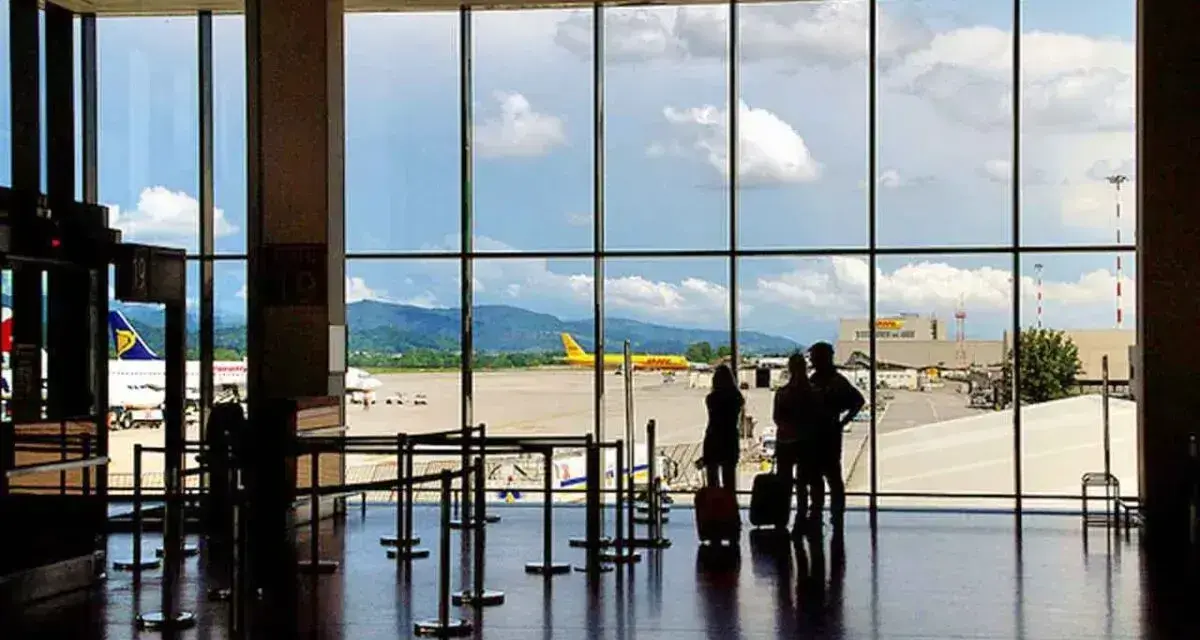Widok z okna na lotnisko Bergamo. Trzy osoby z walizkami czekają na samolot. W tle żółty samolot DHL i góry.