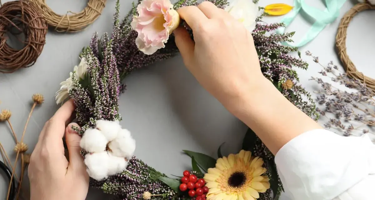 Hände gestalten einen Blumenkranz mit Heidekraut, Gerbera und Baumwolle. Das Binden eines Kranzes ist eine kreative Auszeit.
