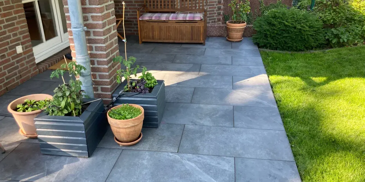 Moderne Terrassenplatten grenzen an einen gepflegten Rasen. Pflanzen in Töpfen und ein Holzbänkchen bieten schöne Rasenkante Ideen.
