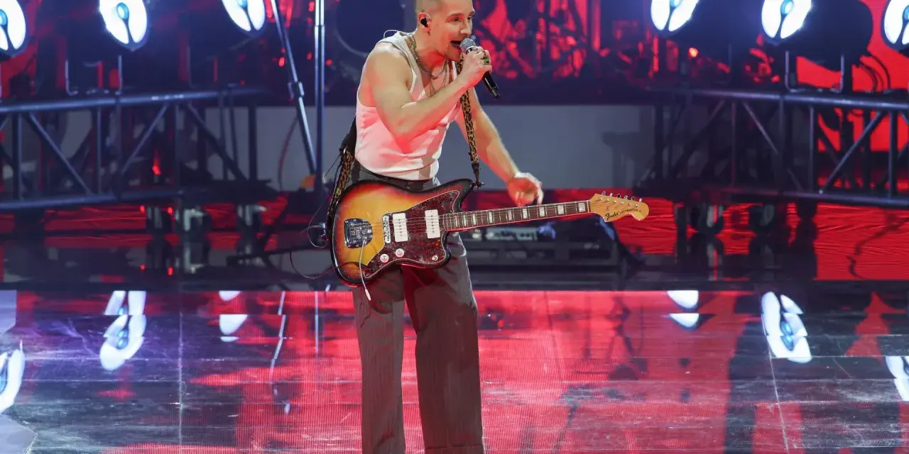 Krzysztof Zalewski na koncercie, śpiewa i gra na gitarze. Scena oświetlona czerwonymi światłami.