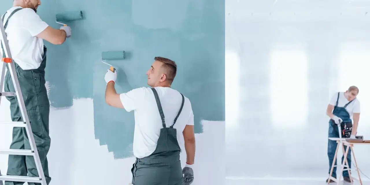 Malowanie ścian na miętowo. Dwóch fachowców na drabinie i stojącego maluje, trzeci pracuje przy stole. Sprawdź, ile za metr malowania ścian!