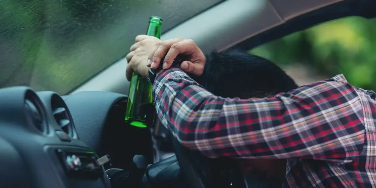Mężczyzna w kratę, z butelką w ręku, opiera się o kierownicę. Pytanie: po ilu latach występuje przedawnienie za jazdę pod wpływem alkoholu?