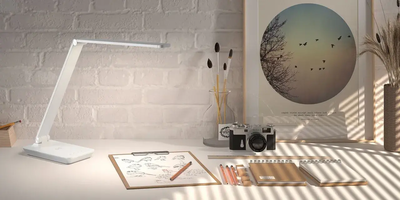 Lámpara LED moderna sobre un escritorio con bocetos de pájaros, cámara antigua y un póster inspirador. La luz es ideal para estudiar.