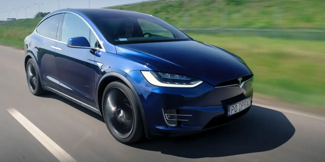 Granatowy Tesla Model X w ruchu na drodze, z widocznym kierowcą.