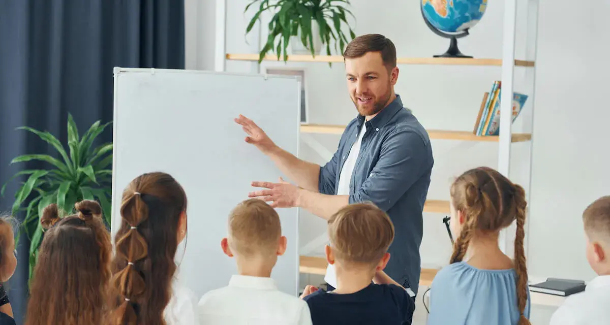 Nauczyciel tłumaczy coś dzieciom przy tablicy. To mogą być dni dyrektorskie, podczas których uczniowie poznają szkołę.