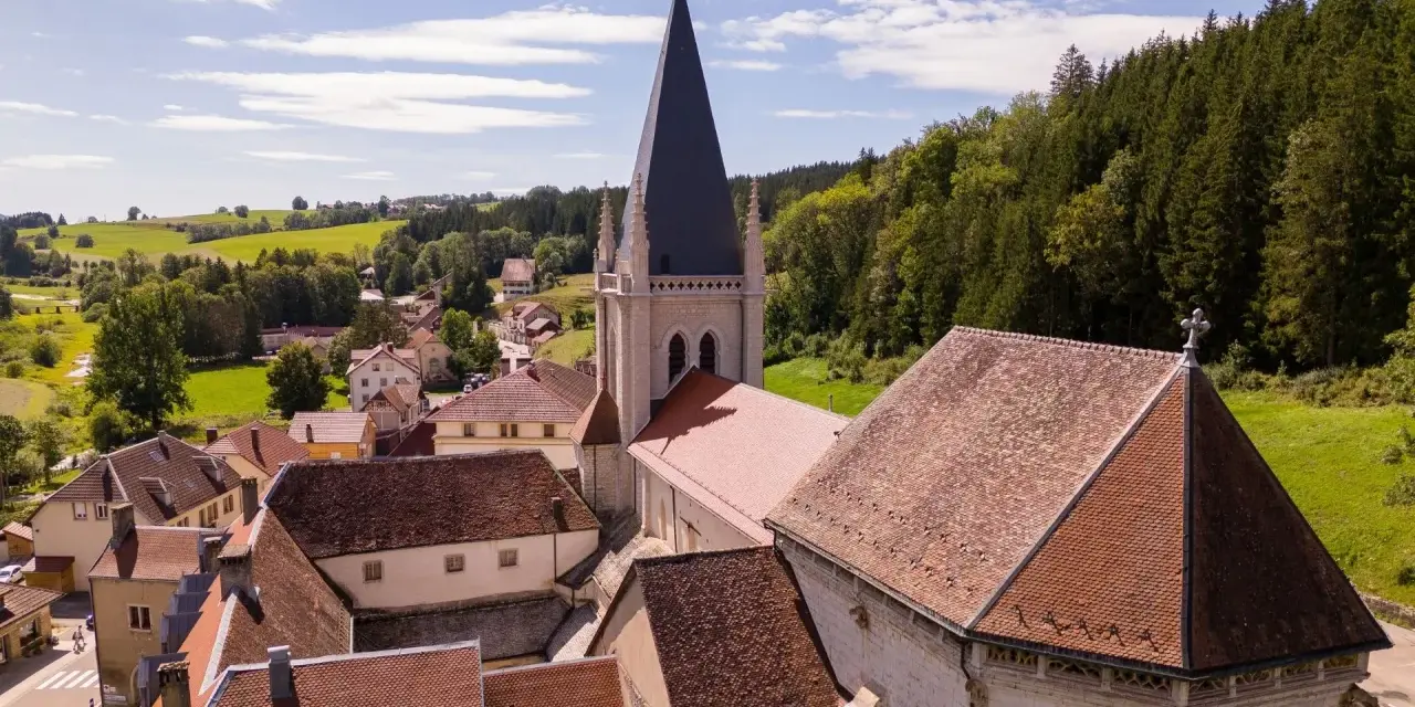 Vue aérienne d'un village pittoresque du haut Doubs, dominé par une église à la flèche élancée, entouré de collines verdoyantes et de forêts.