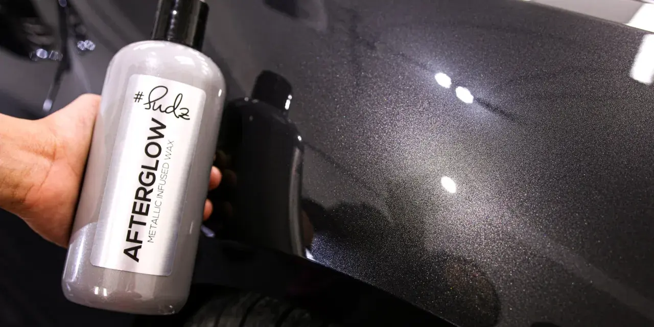 Bottle of "Afterglow Metallic Infused Wax" held against a dark, metallic car finish, showcasing the product's ability to create multiple coats of wax for a deep shine.