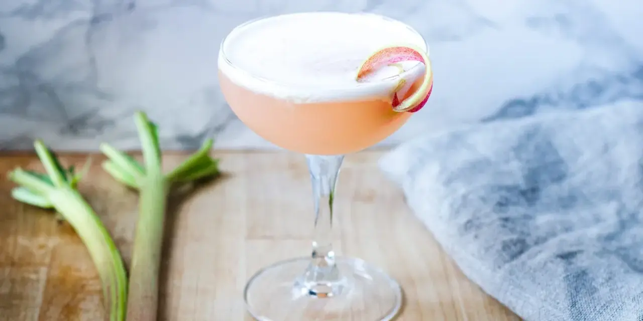 En rosa rabarber sour-cocktail med skum och en rabarberdekoration. Två rabarberstjälkar ligger bredvid.