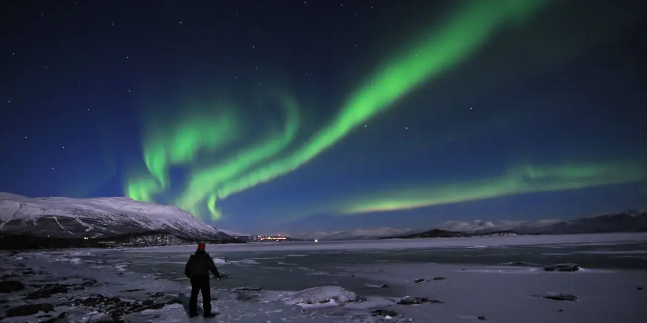 En ensam person står på en isig strand och ser på det magiska abisko norrsken som dansar över himlen.