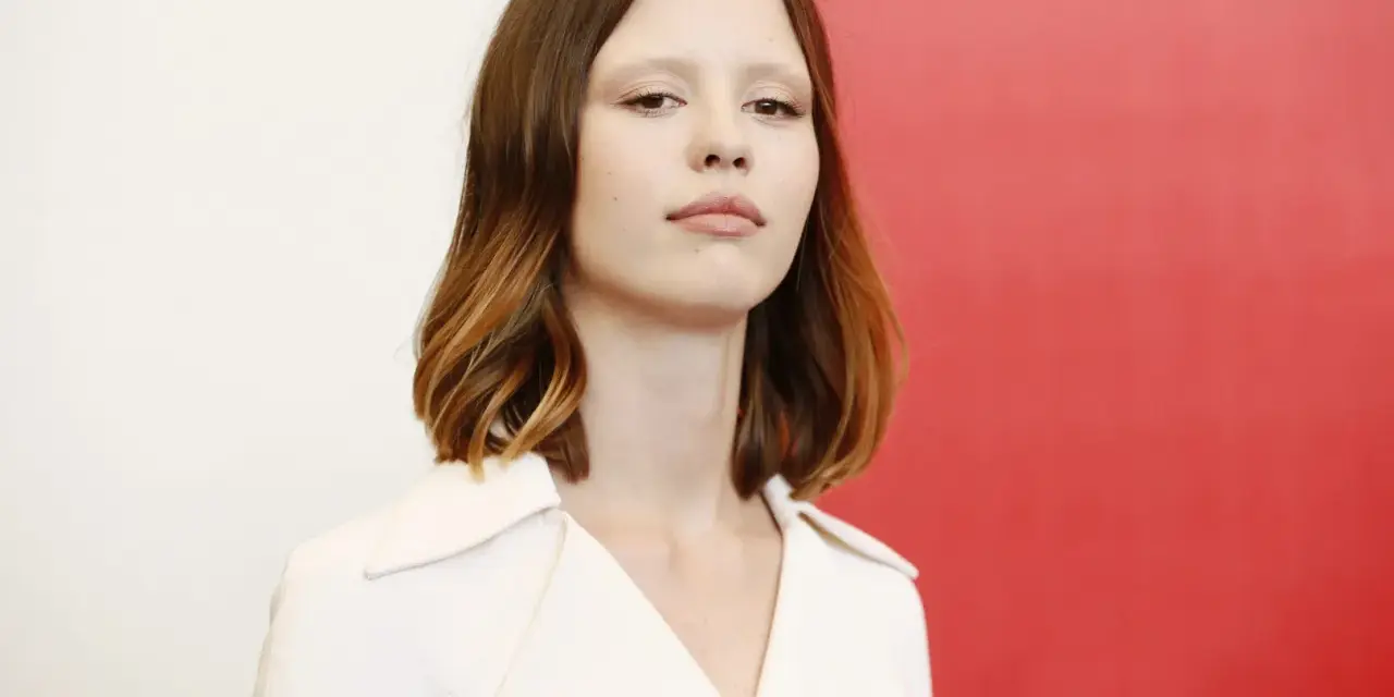 La giovane attrice protagonista del film di mia goth, con capelli castani mossi e un'espressione intensa, indossa un abito bianco.