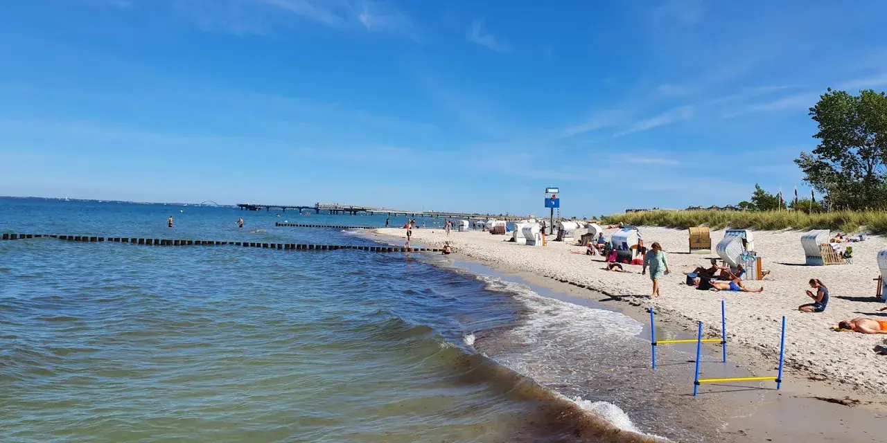 En solig tysk strand med folk som njuter av havet och sanden. En brygga sträcker sig ut i vattnet.