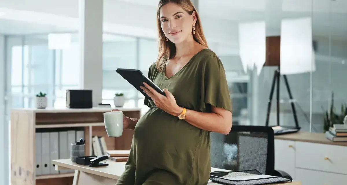 Uśmiechnięta ciężarna kobieta z tabletem i kubkiem w biurze. Dowiedziałam się o ciąży po okresie wypowiedzenia, ale teraz planuję przyszłość.