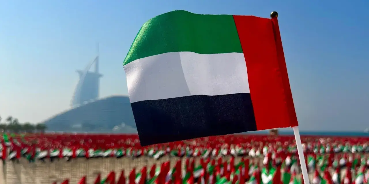 Flaga Zjednoczonych Emiratów Arabskich powiewa na tle drapaczy chmur w Dubaju. W tle widać morze małych flag.