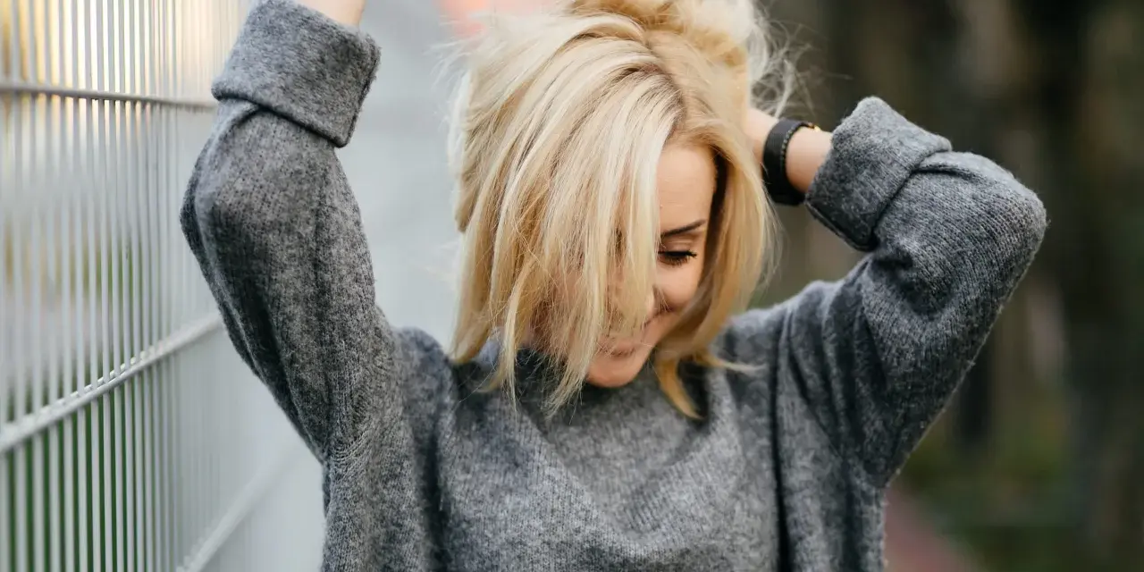 Blondynka w szarym swetrze, z włosami opadającymi na twarz. Marzy o gładkiej skórze, rozważa depilację laserową jasnych włosów.